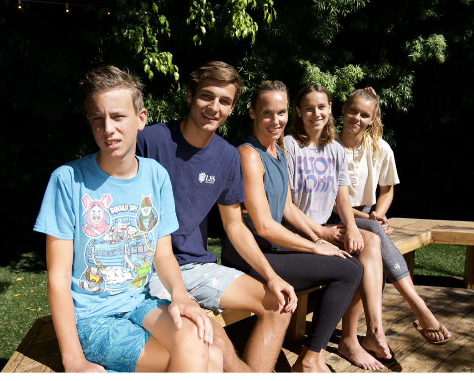 Five people sit closely on a wooden bench outdoors, smiling at the camera with trees in the background on a sunny day.
