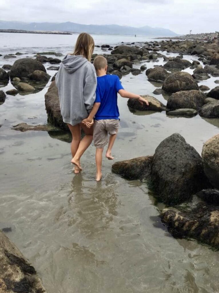 Two children walk hand in hand along a rocky shoreline, wading through shallow water at the beach.