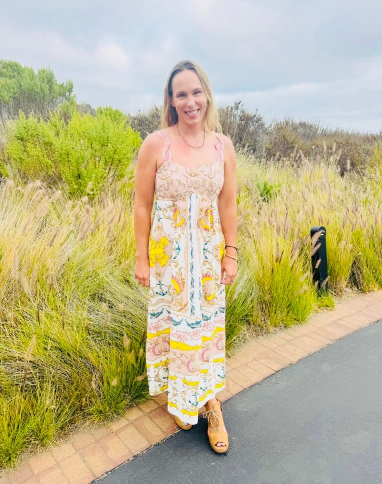Smiling woman in a colorful patterned maxi dress standing on a paved path with tall grasses and cloudy sky behind her.